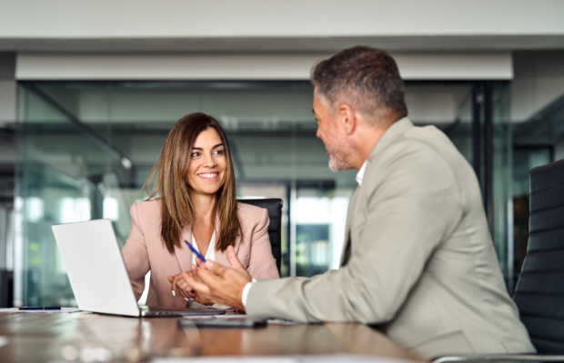Happy professional mature Latin businessman and businesswoman executive colleagues wearing suits sitting at table having partnership business discussion using laptop at corporate meeting in office.